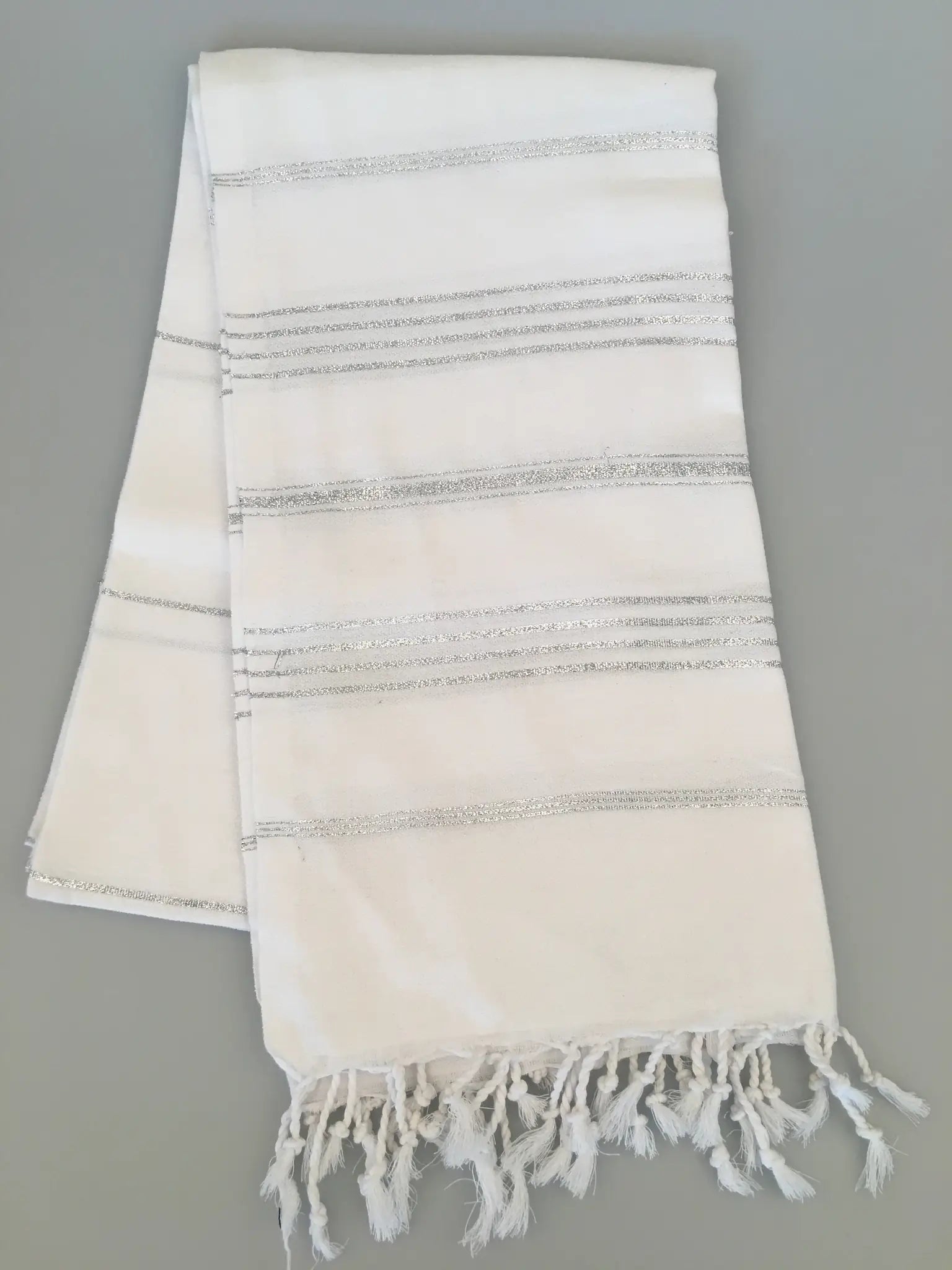 White Turkish towel with silver striped patterns and fringed edges, laid flat on a light gray background.