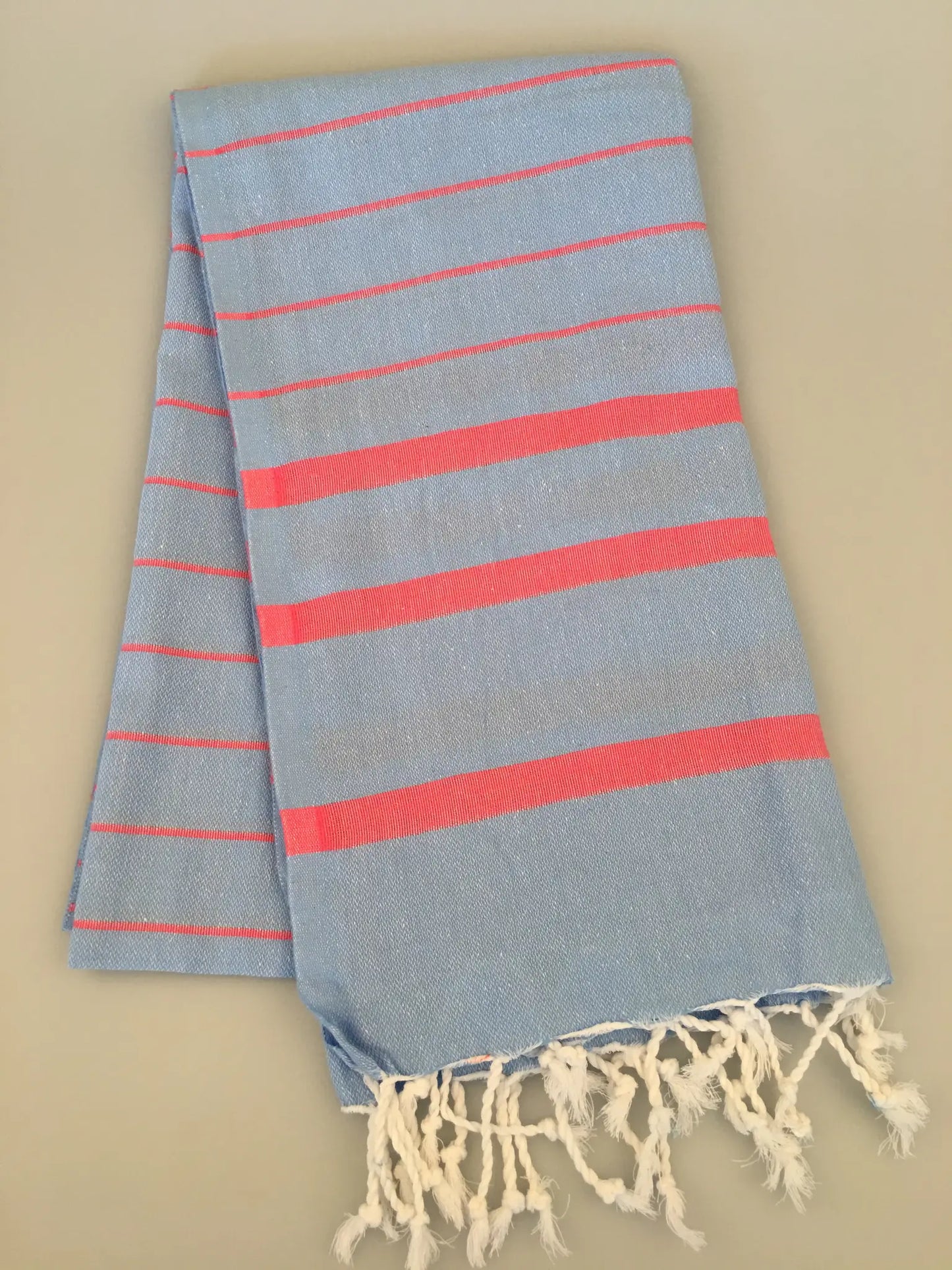 Light blue Turkish towel with pink stripes and fringed edges, laid flat on a light beige background.