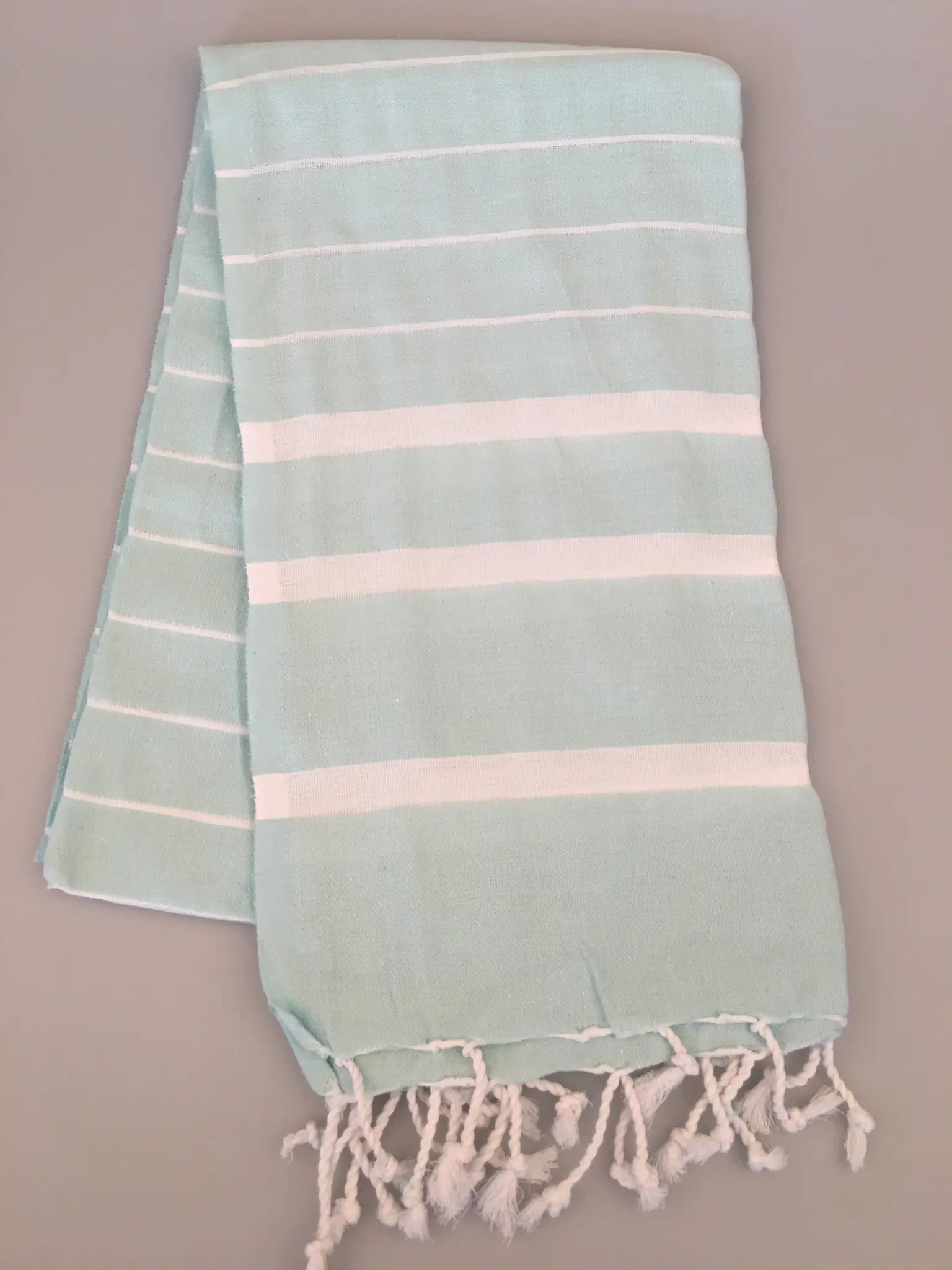 Light blue Turkish towel peshtemal with white stripes and fringed edges, displayed flat on a gray background.