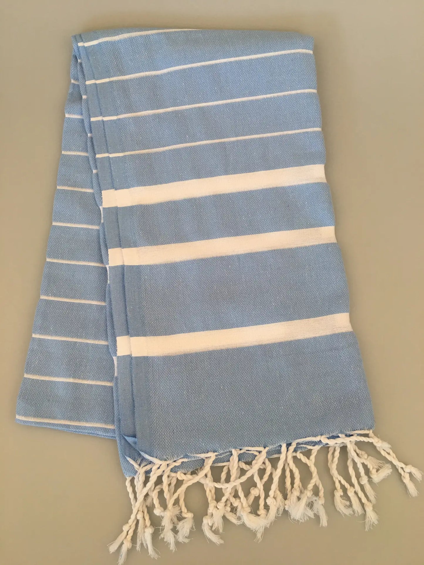Light blue Turkish towel peshtemal with white stripes, featuring fringed edges, laid out flat on a neutral background.