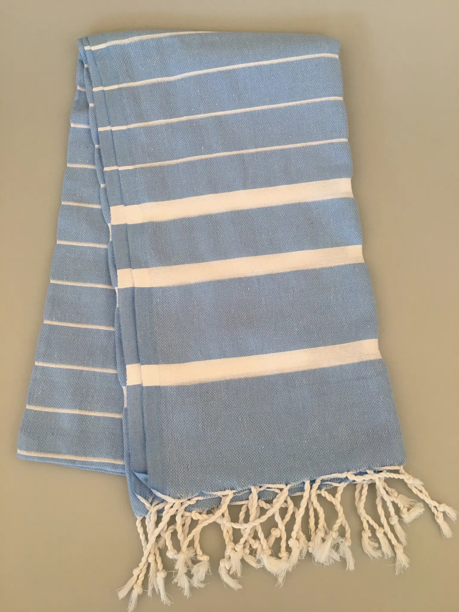 Light blue Turkish towel peshtemal with white stripes, featuring fringed edges, laid out flat on a neutral background.
