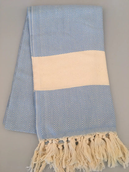 Light blue Turkish towel peshtemal with herringbone pattern, cream stripe, and fringed edges, folded on a gray background.