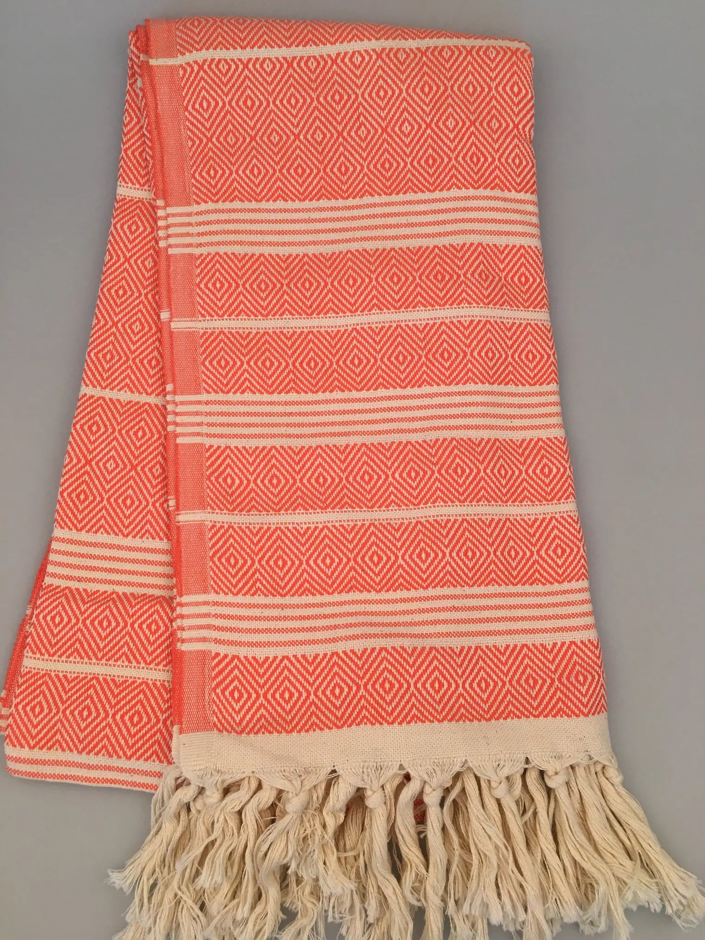 "Coral and cream Turkish towel with diamond pattern and fringed edges, folded on a gray background."