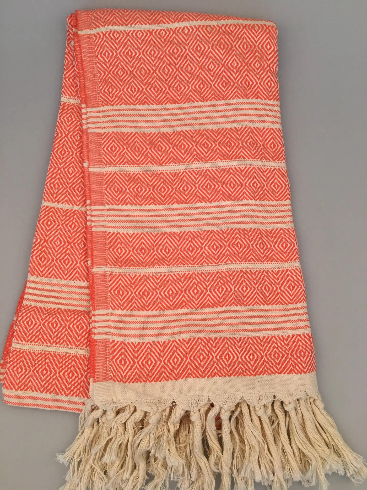 "Coral and cream Turkish towel with diamond pattern and fringed edges, folded on a gray background."