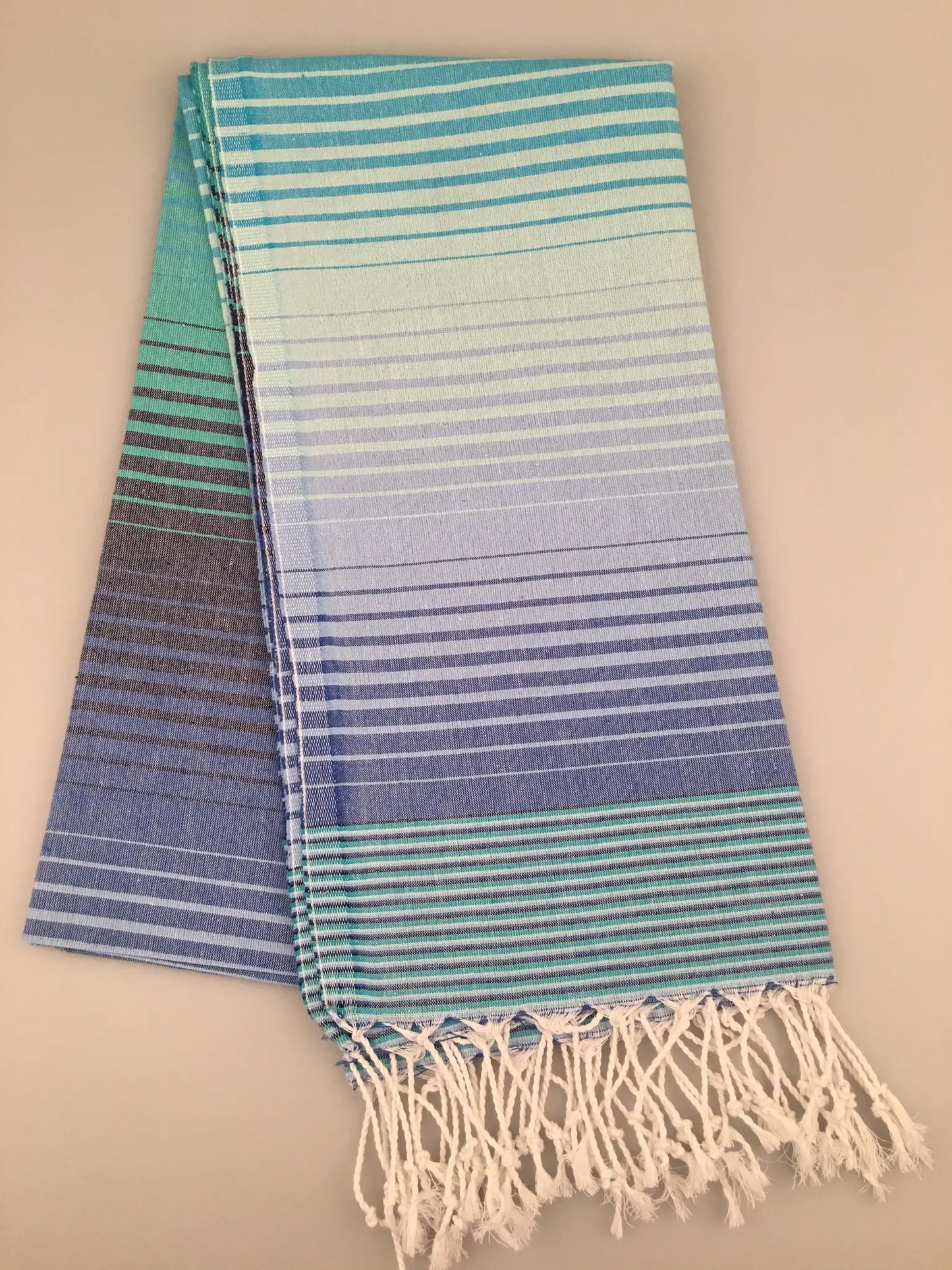 "Light blue Turkish towel with gradient stripes, fringed edges, folded view on neutral background."