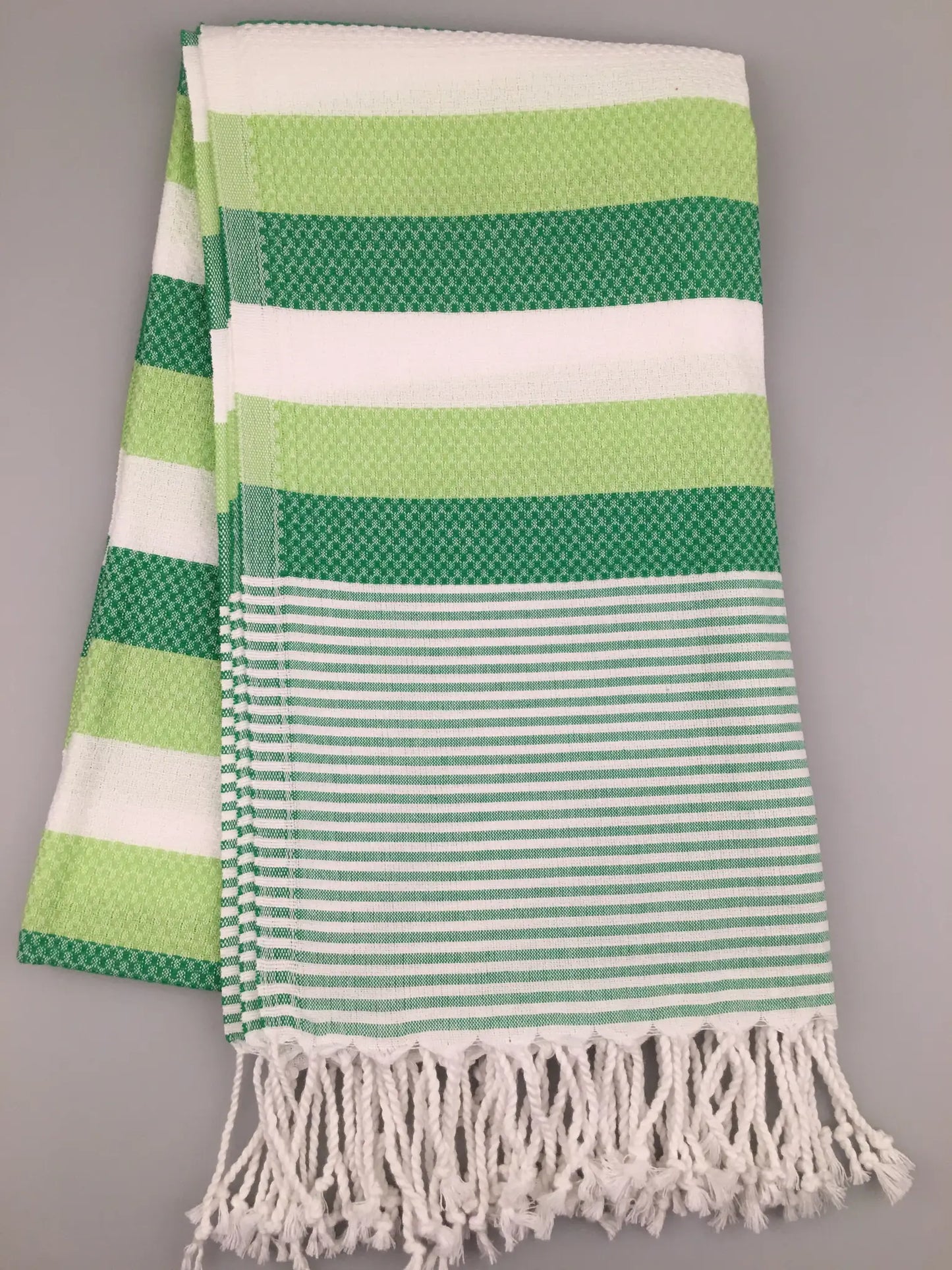 Green and white striped Turkish towel with textured patterns, featuring fringed edges, laid flat on a gray background.