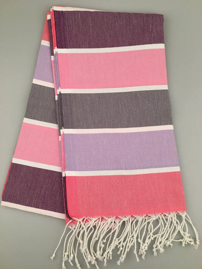 "Tripolis Turkish Towel Peshtemal in pink, purple, and gray stripes, folded with white fringe at the ends."