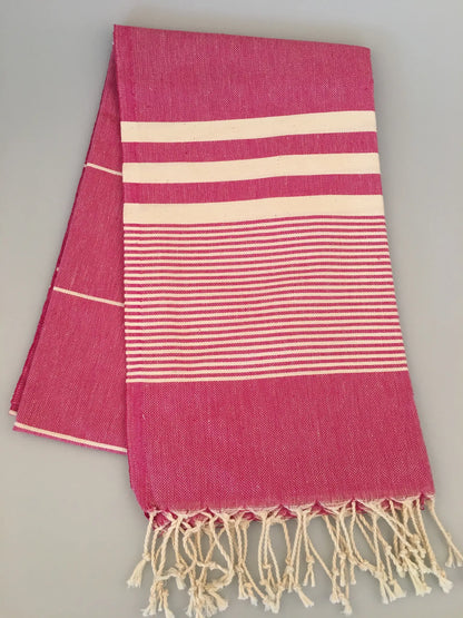 "Pink Turkish towel with white stripes and fringed edges, laid flat on a light gray background."