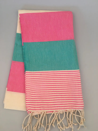 "Turquoise and pink striped Lagina Turkish towel with cream accents and fringed edges, folded on a gray background."