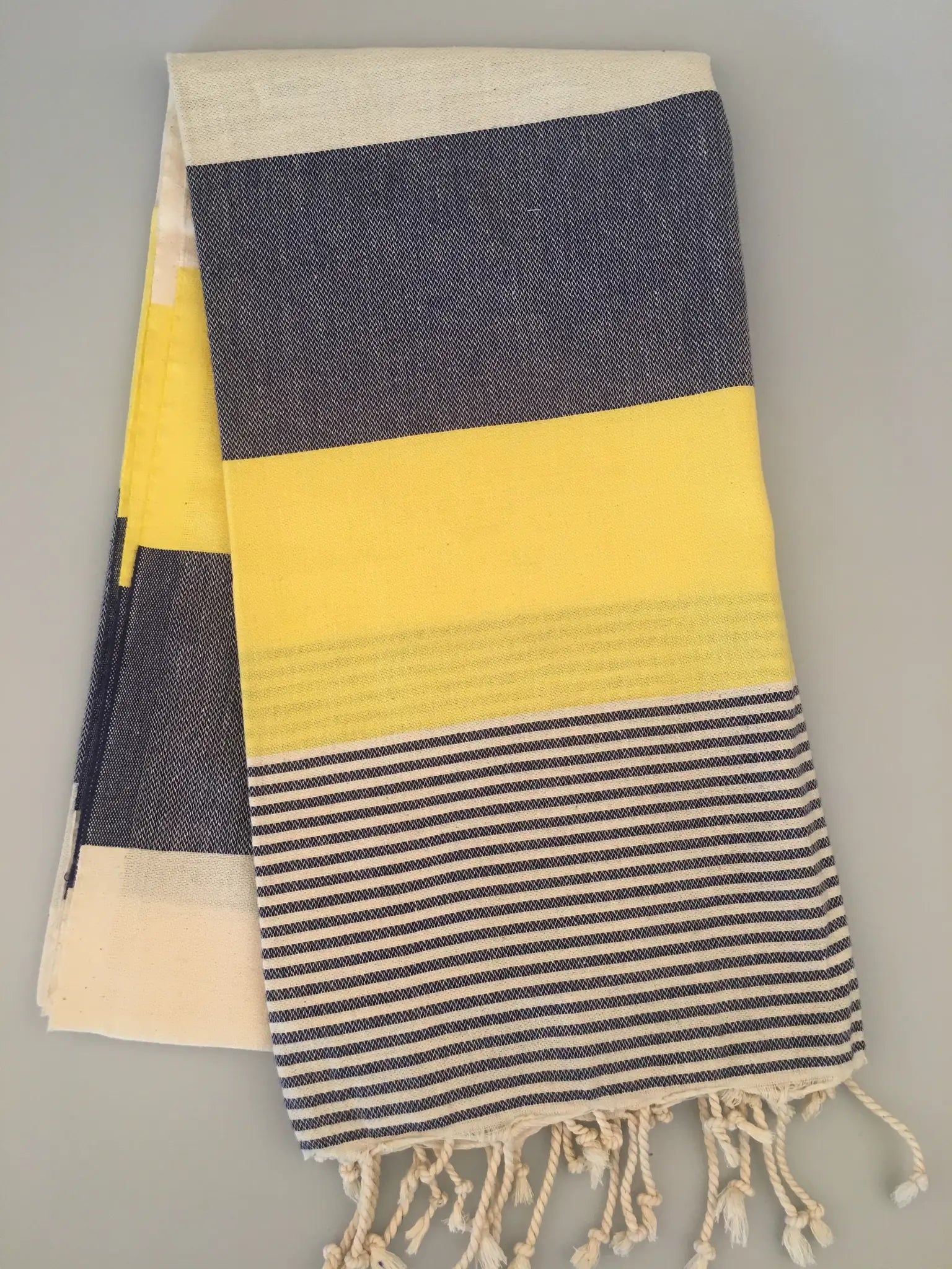 "Yellow and navy striped Turkish towel with fringed edges, folded on a light gray surface."