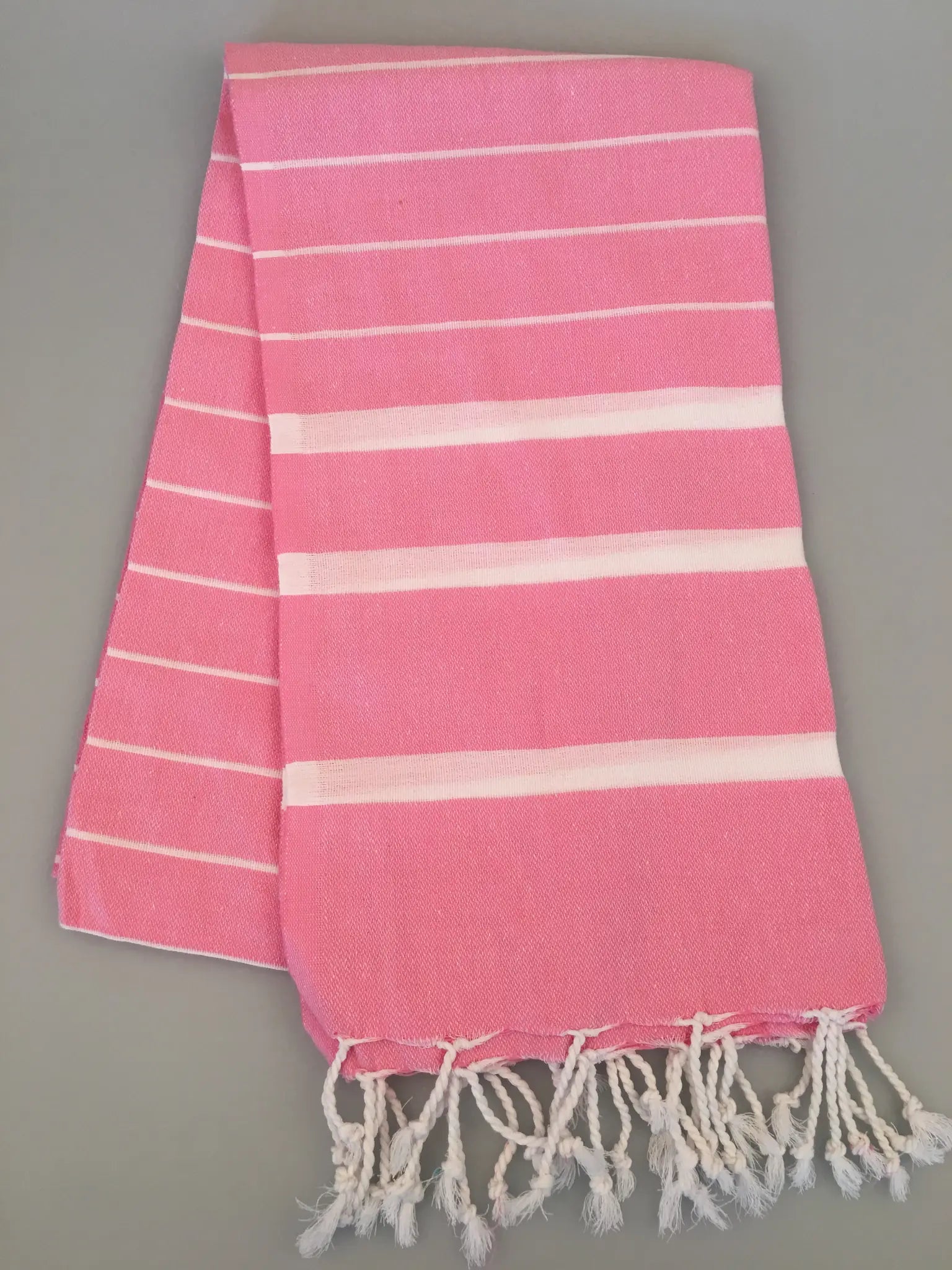"Pink Turkish towel peshtemal with white stripes and fringe edges, laid flat on a gray background."